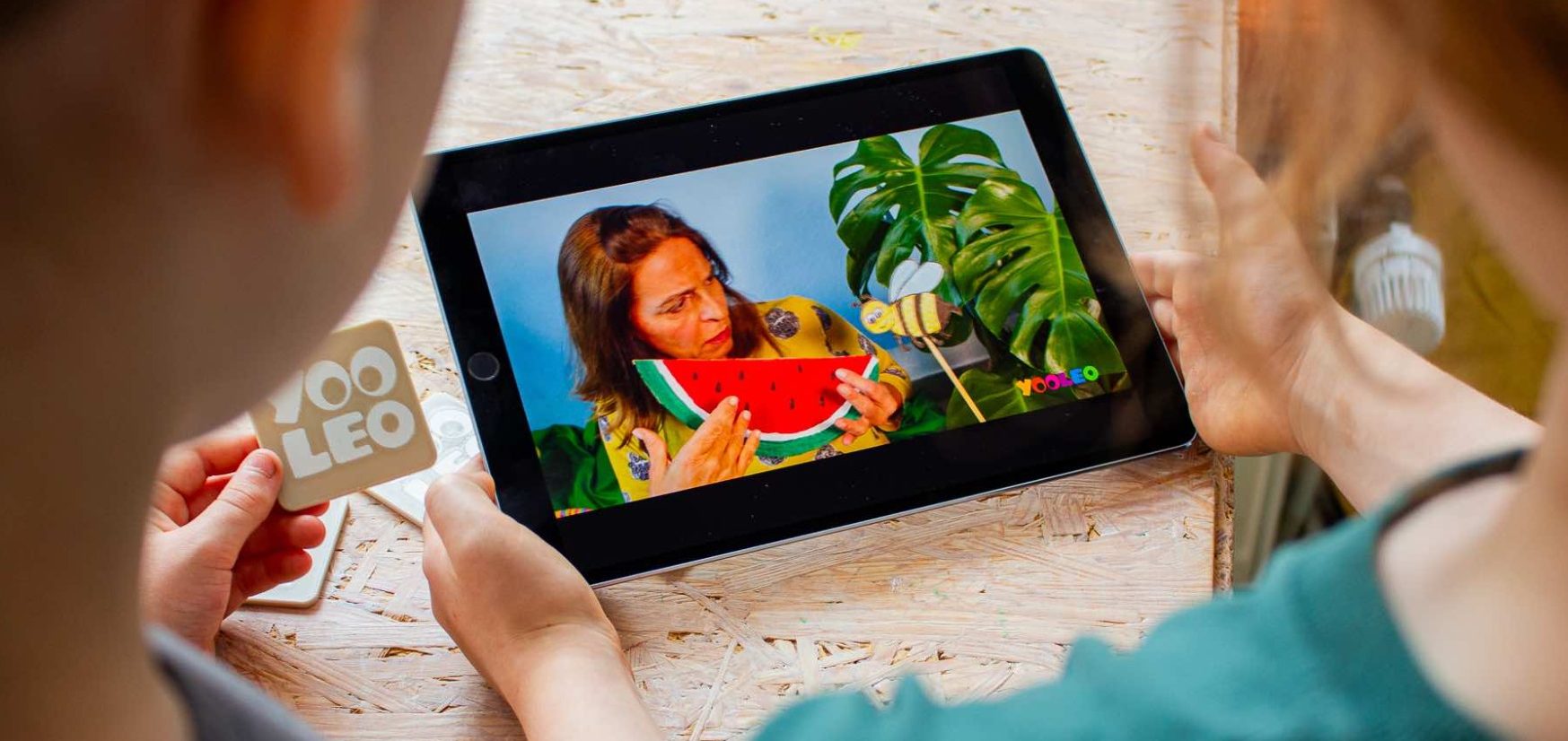 Zwei Kinder schauen gemeinsam ein Video auf einem Tablet. Im Video ist eine Frau mit Melone und Pflanzen zu sehen.