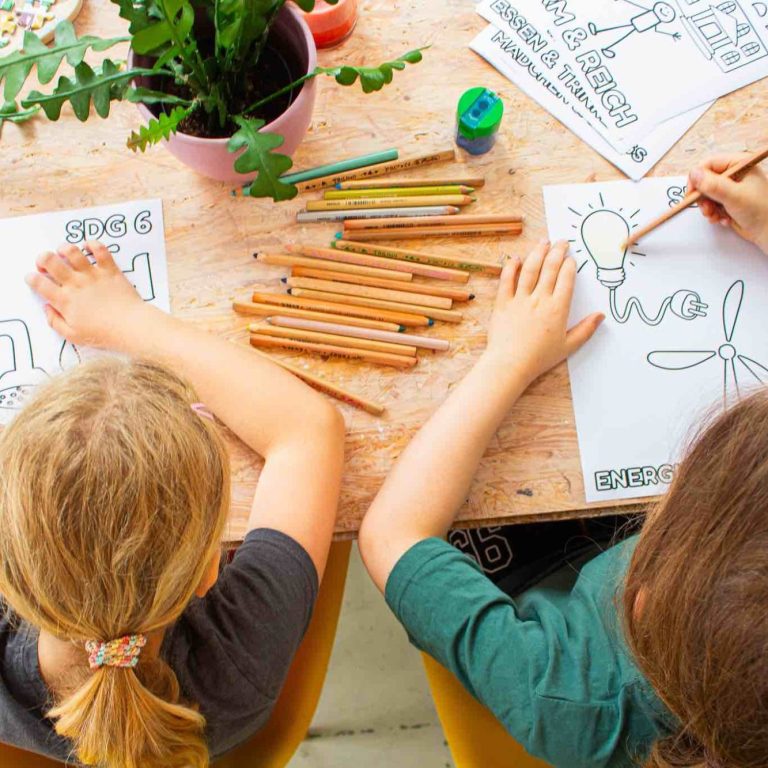 Zwei Kinder zeichnen auf Papier, umgeben von Bastelmaterial und einer Pflanze.